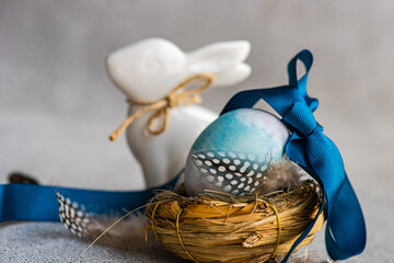 A close up Easter composition with a blue dyed egg adorned with a ribbon in a nest, guinea fowl feather, and a ceramic rabbit on a textured background