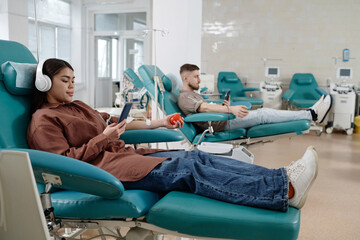 Obraz premium Blood donors sitting side by side on medical recliner chairs in hospital and browsing smartphones