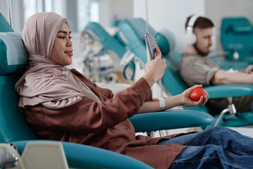 Muslim woman taking selfie during blood donation process in clinic, young man sitting on background - obrazy, fototapety, plakaty