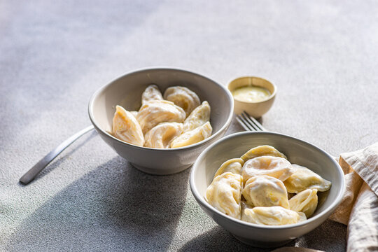 A pair of ceramic bowls containing Ukrainian varenyky, traditional dumplings, served with a side of sour cream and butter sauce, illuminated by natural light