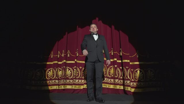 Male entertainer in a suit enters the stage opening a red curtain. The artist on stage in the light of the soffit spotlight.