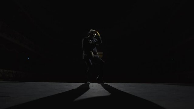 Adult man in a suit dances on a dark stage of a theater in the glow of a spotlight. Silhouette of a dancer in a hat.