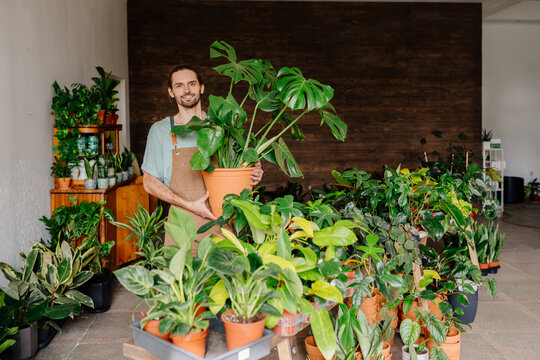 Portrait Of A Young Asian Woman Business Owner Holding Many House Plants Smiling And Looking At Camera In A Greenhouse. Gardener Working In Flower Shop, Plant Store.