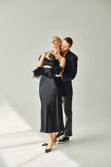 appealing sexy couple in elegant black attires posing together and hugging lovingly on gray backdrop