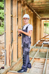 Carpenter constructing wooden framed house. Man worker in glasses working with screwdriver, wearing work overalls and helmet. Concept of modern eco-friendly construction.