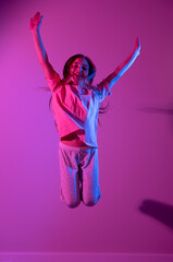 Portrait of cute smiling girl jumping with hands up on pink background with blue neon light. Party concept. Different emotions. Happiness, childhood, holidays, women's day, lifestyle. Copy space. 