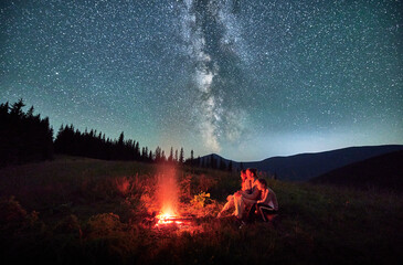 Cozy night with family in mountains. Young family sitting next to bonfire at starry night. Bright view of Milky Way over hills. - obrazy, fototapety, plakaty