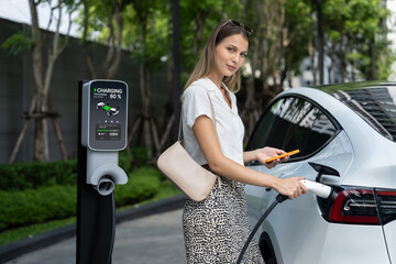 Young woman travel with EV electric car charging in green sustainable city outdoor garden in...