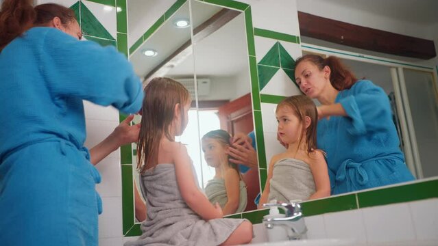 Happy mother combing her little cute daughter's hair in front of the mirror after taking a bath and washing her hair. Time spent with your child. Child care, motherhood