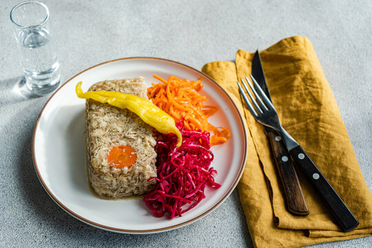 Top view of slice of chicken aspic with a carrot centerpiece, served with spicy shredded carrot and pickled beetroot cabbage, accompanied by a shot of vodka