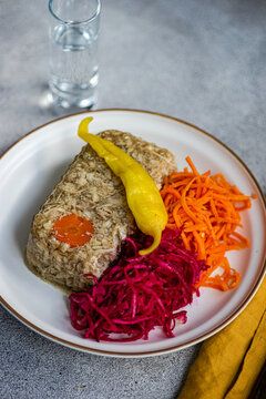 Top view of chicken aspic garnished with a carrot slice, accompanied by spicy shredded carrot and pickled beetroot cabbage salad, presented with a clear glass of vodka on a white plate