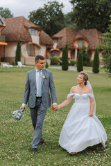 Fototapeta na wymiar A handsome groom and an elegant bride in a lush white dress are walking in a summer park. Happy bride and groom getting ready for their best day.