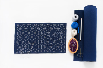 Process of hand embroidery in  Sashiko style on blue canvas with white threads of geometric pattern, accessories, pincushion, needles, scissors, threads, canvas on white table. Flat lay, copy space