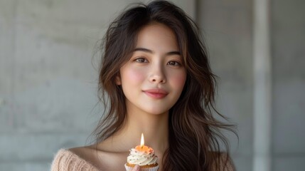 a woman holding a cupcake with a candle in her hand and looking at the camera with a smile on her face.