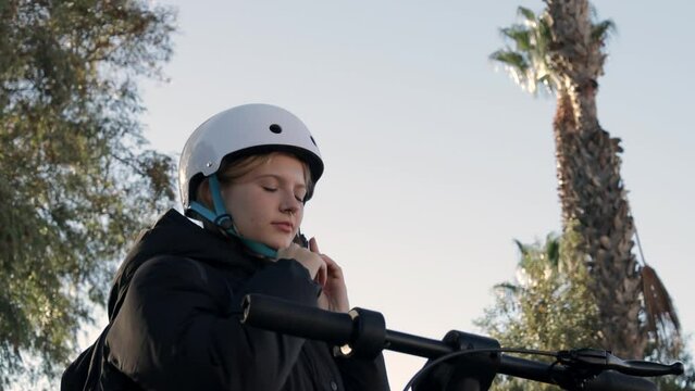 A young girl puts on a white helmet while standing near an electric bicycle on a palm alley,blue sky