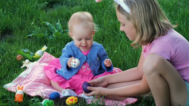 Cute baby girl with her older sister having egg tapping while sitting on the grass outdoors, slow motion. Easter hunt concept