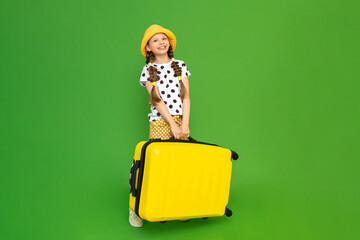 A little girl is holding a big heavy suitcase for trips. Vacation for teenagers at a summer camp. A happy child in a hat and shorts is preparing to travel abroad. Green isolated background.