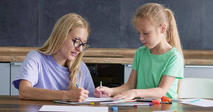 A young girl with glasses draws with pencils together with a child, the teacher teaches the girl to draw on paper.