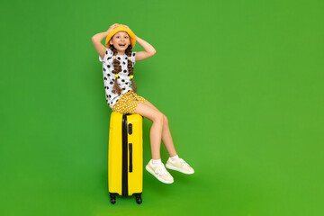 A little girl is sitting on a yellow suitcase and is happy about the upcoming trip to a summer children's camp. A child in a hat and shorts is smiling happily. Green isolated background.