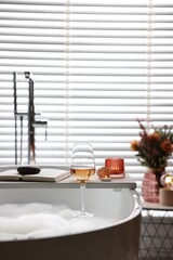 White wooden tray with glass of rose wine, book and burning candles on bathtub in bathroom