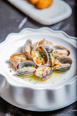 Almejas a la marinera, decoradas con cebollino y salsa de vino albariño en un plato blanco sobre una mesa de marmol negro acompañadas de coquetas de marisco