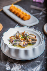 Almejas a la marinera, decoradas con cebollino y salsa de vino albariño en un plato blanco sobre una mesa de marmol negro acompañadas de coquetas de marisco