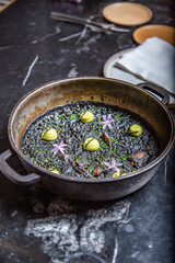 Arroz negro de sepia con alioli de algas codium, flores decorativas, en una paella de hierro fundido sobre una mesa de marmol negro de un restaurante con clase