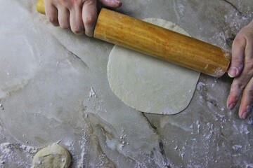 on the kitchen table, women's hands roll out dough with a rolling pin. several round pieces of dough and a prosky piece for stuffing it with meat are visible.
