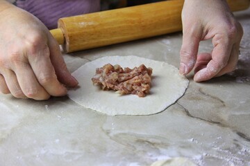 on the  table there is a flat pancake made of raw dough and in the center there is a pile of minced beef with onions. female hands wrap the edges for steaming manti dumplings. cooking homemade food