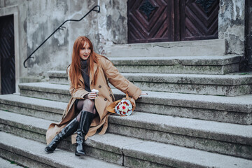 A trendy ginger woman is having coffee to go while sitting on rustic stairs.
