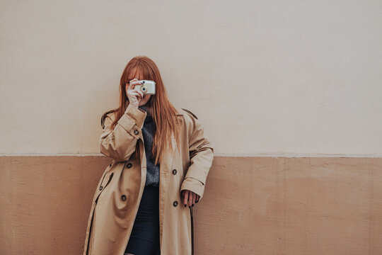 A trendy lady is taking pictures with her camera while leaning on a wall.