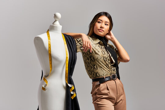 creative asian designer looking away near mannequin with fabric and measuring tape on grey