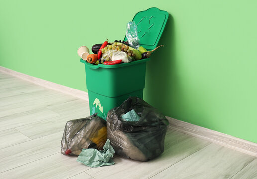 Container for trash with food waste and full garbage bags near green wall. Recycling concept