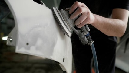 A worker professionally cleans the surface of a car bumper with a grinding machine in a car service garage before painting, close-up. Sanding a car bumper before painting.