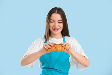 Pretty young woman in apron with fresh croissant on blue background