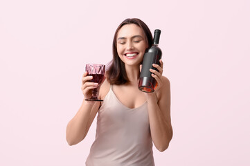 Beautiful young woman with glass and bottle of red wine on pink background