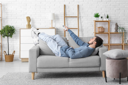 Handsome young man with air conditioner remote control lying on sofa in living room