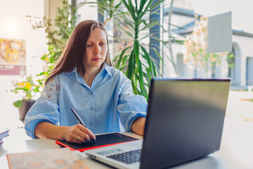 Young female teacher getting ready for lesson working in office using laptop. Woman writing notes...