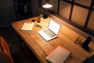 Naklejka premium Blank laptop with glowing lamp on desk in dark office at night