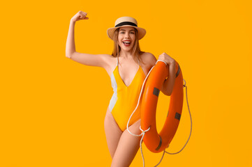Pretty young woman in tank swimsuit and with lifebuoy on yellow background