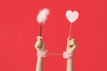 Female hands with handcuffs, feather stick and paper heart on red background. Valentine's Day...
