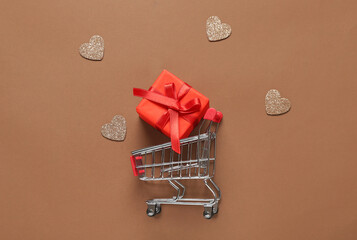 Small shopping cart with gift box and hearts on brown background. Valentine's Day celebration