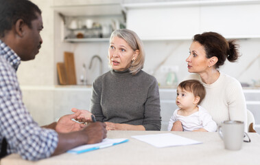Obraz premium Two thoughtful women holding little baby sitting at the kitchen-table face to face with male agent demonstrating some documents