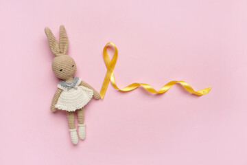 Golden ribbon with toy bunny on pink background. Childhood cancer awareness concept - obrazy, fototapety, plakaty