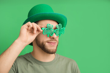Young man in leprechaun hat and decorative glasses in shape of clover on green background. St....