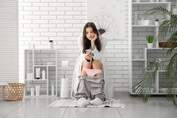 Young woman using mobile phone on toilet bowl in restroom - obrazy, fototapety, plakaty