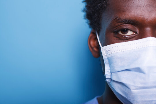 Close-up of the eye of African American person wearing a face mask and staring at camera. Portrait of man having coronavirus epidemic protection for healthcare, standing over blue background.