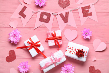 Garland made of paper flags with word LOVE, gift boxes and chrysanthemum flowers on pink wooden background. Valentine's Day celebration