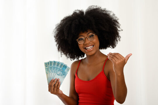 Smiling young black woman holding Brazilian banknotes, positively surprised, gesturing with her hand, space for text, person, advertising concept