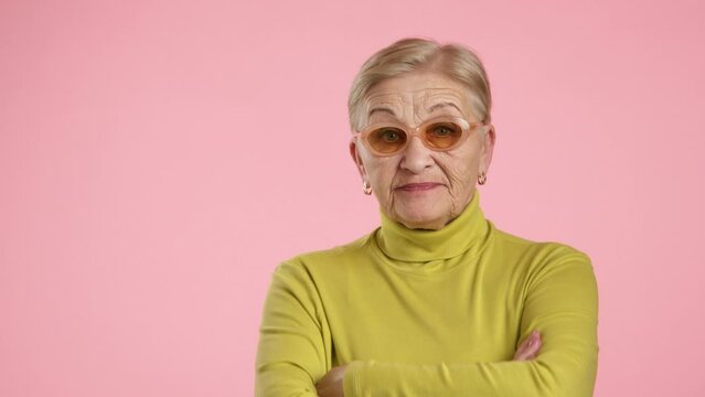 Well-groomed elederly lady with blonde short hair in stylish spectacles, yellow turtleneck standing over pink background, looking with disrespect, contempt. High quality 4k footage